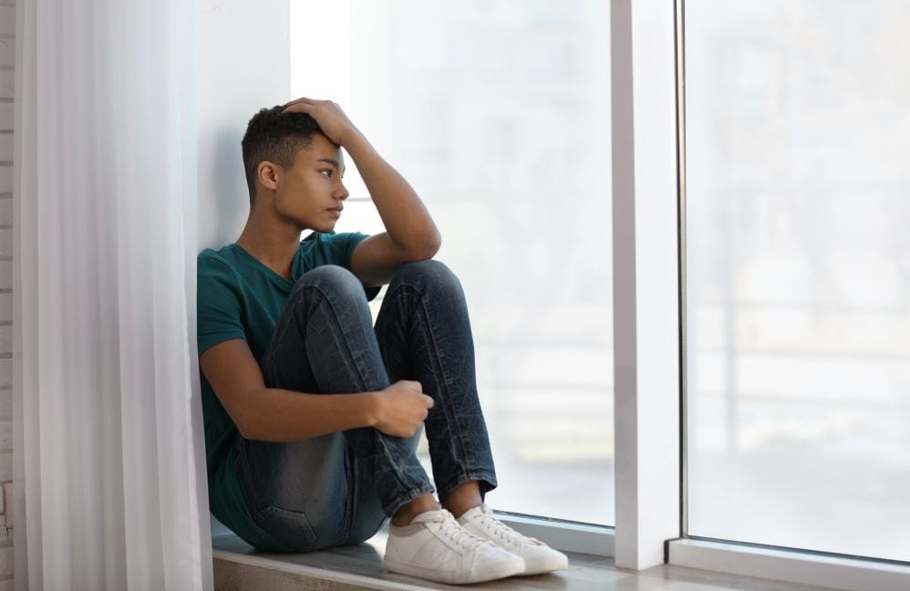 Young teen boy looking out window with hand on his forehead.