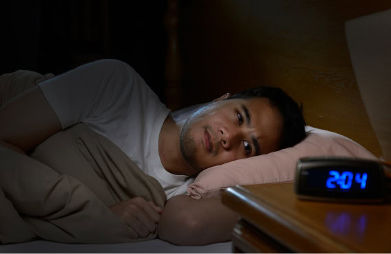 A man laying awake in bed, staring at his alarm clock.