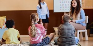 2023-07_website-article-template-570--370-px-1 A group of three children sitting and listening to another student present the alphabet.