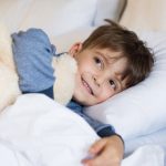 A young boy laying in bed, smiling and holding a teddy bear.